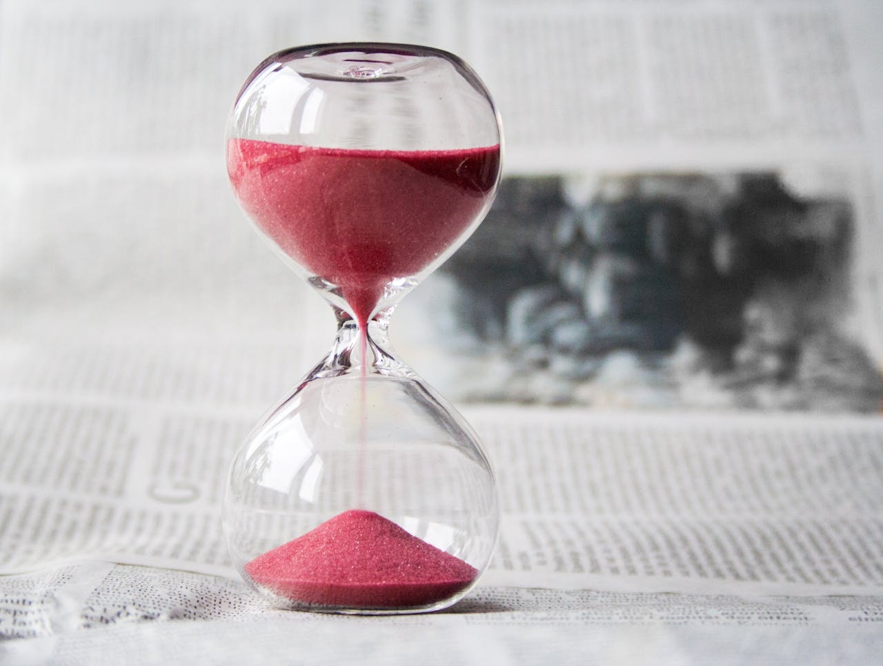 Avasta oma mõtete jõud – 2 nädalat teadlikku fookust Close-up of a transparent hourglass with pink sand flowing, placed on a newspaper background.