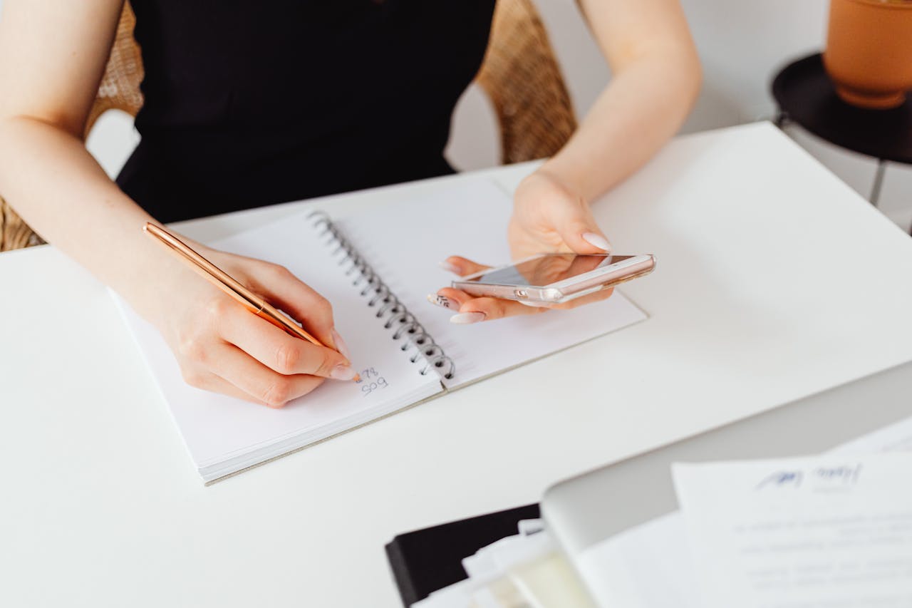 services-04 A woman writes in a notebook while holding a smartphone, symbolizing multitasking at work.